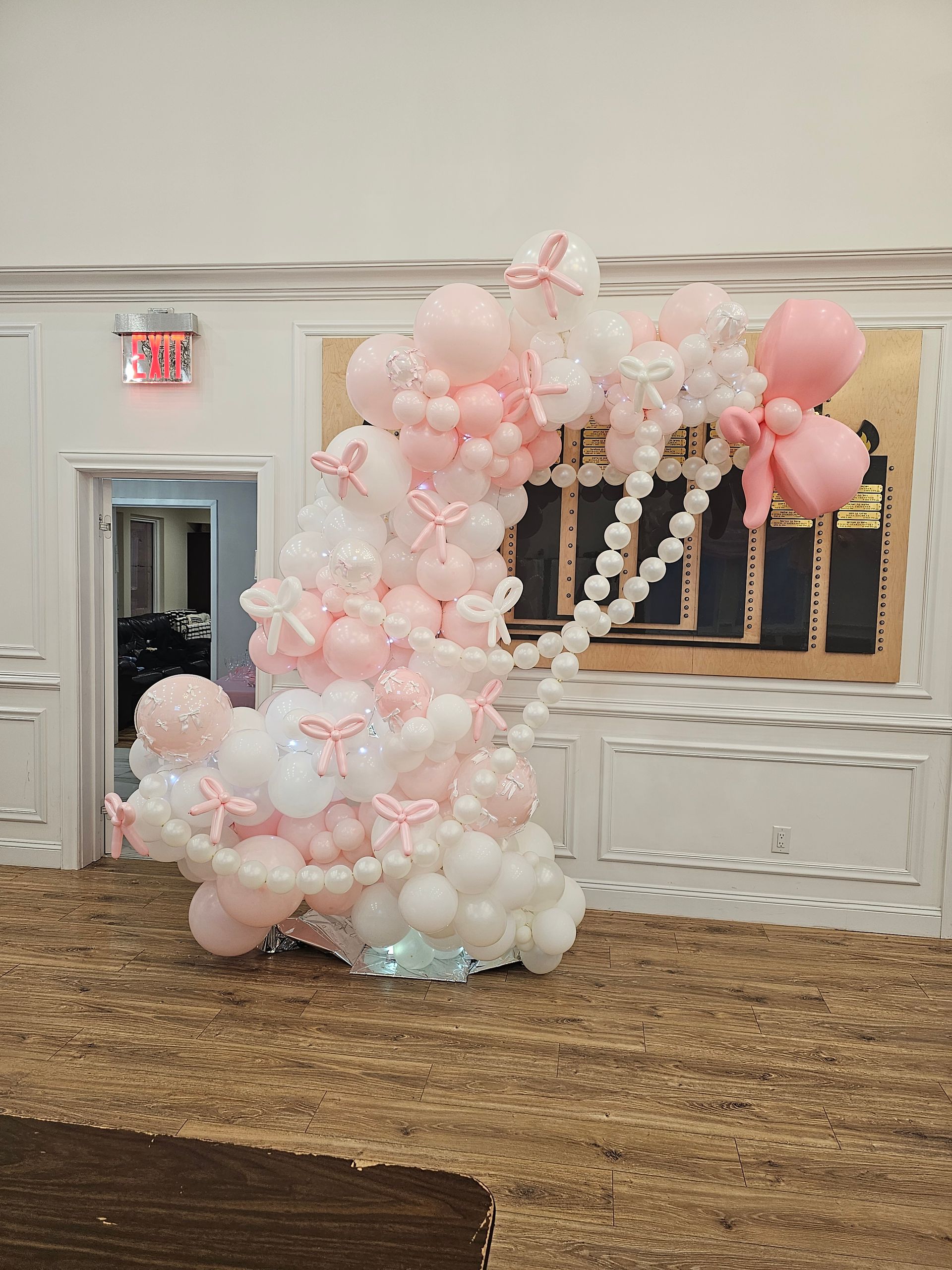 Pink and white balloon arch with bows, in a room with a patterned carpet and gold-toned wall.