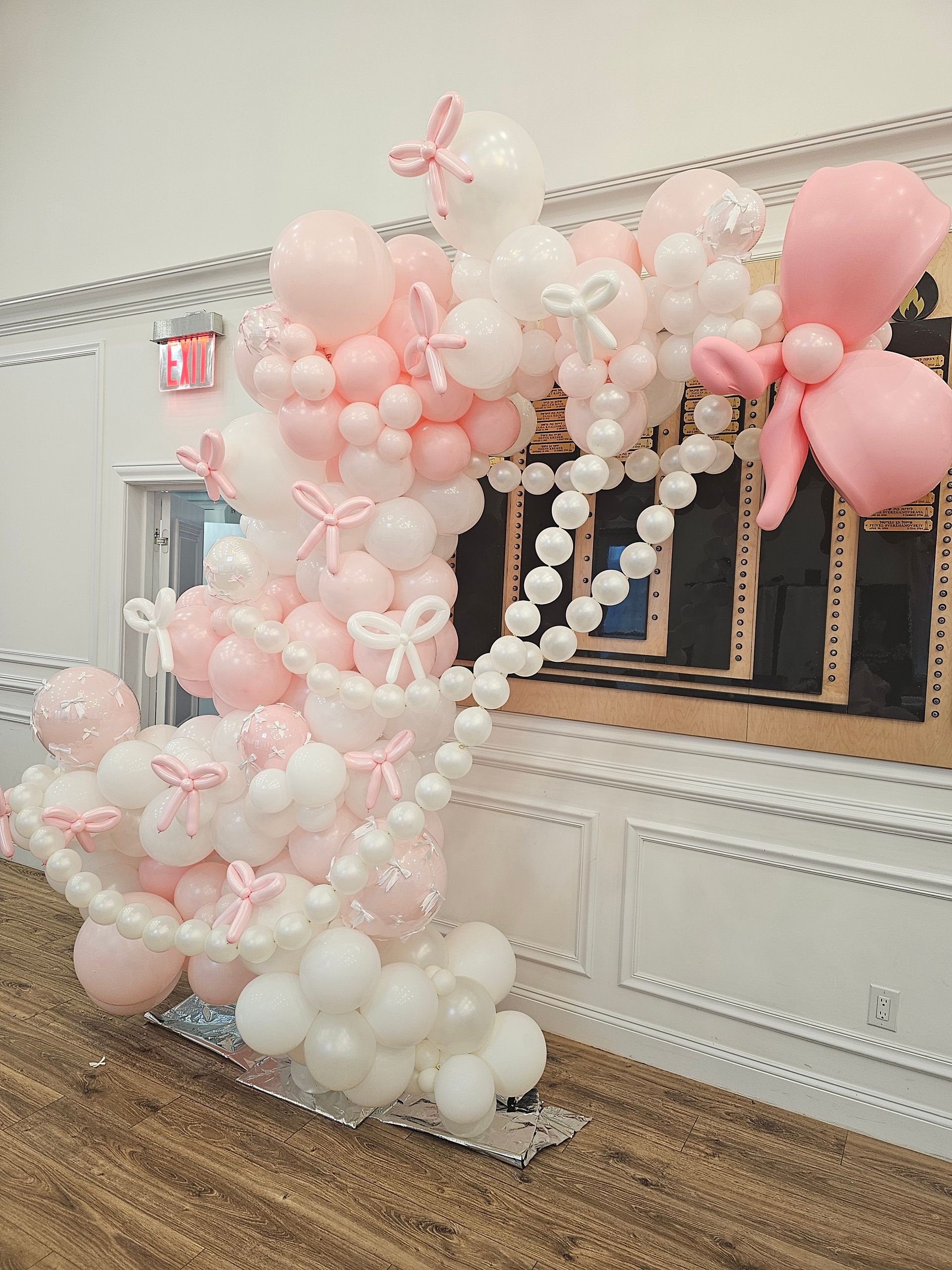 Pink and white balloon arch with bows, against a wall with wainscoting and a mirror base.