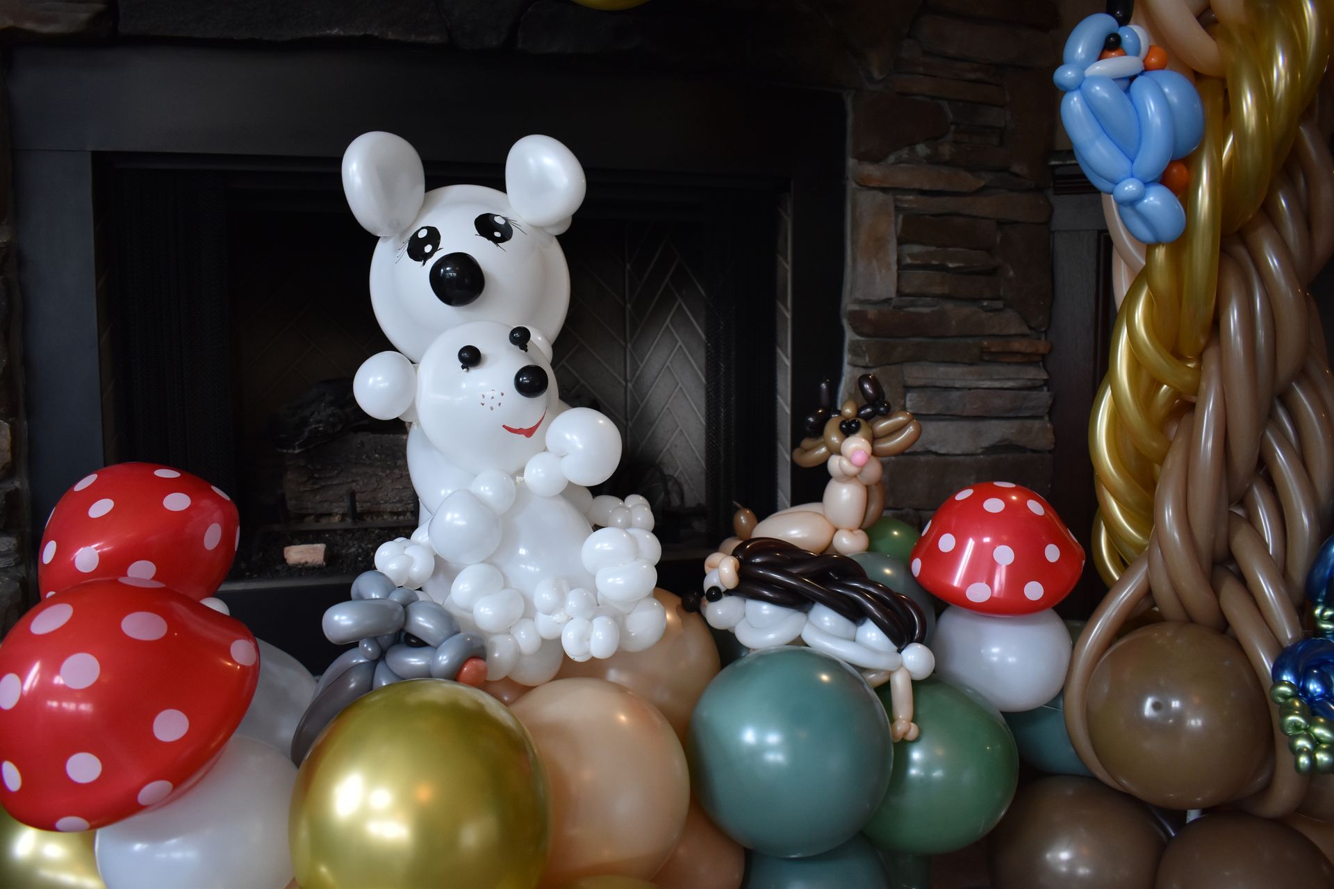 Balloon animals: two mice, a deer, and mushrooms atop a balloon arrangement with a fireplace background.