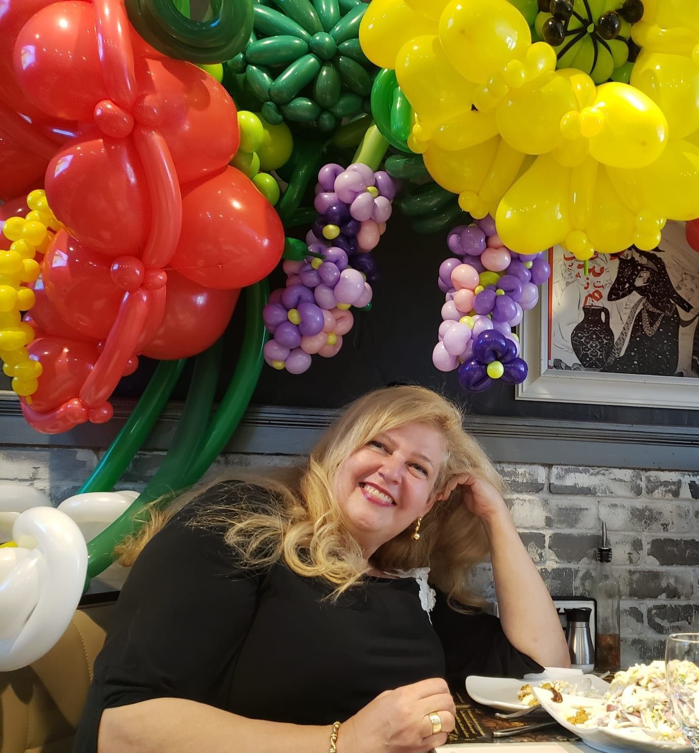 Woman smiling, seated at a table with a colorful balloon fruit arrangement overhead.