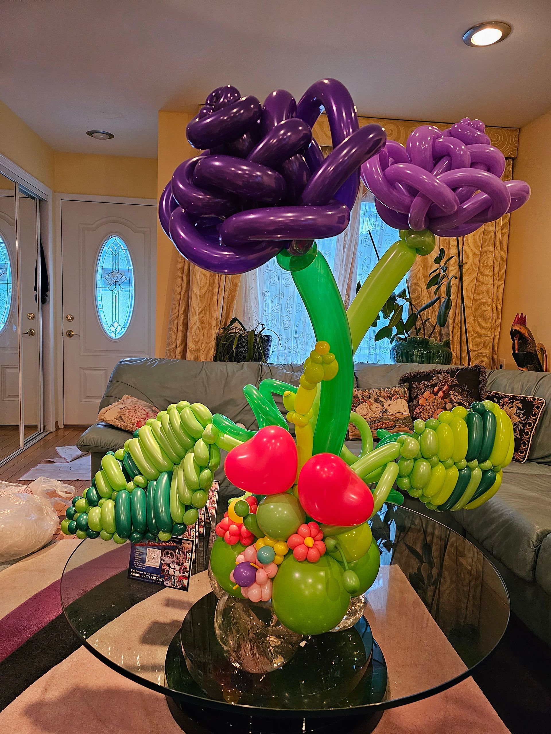 Balloon sculpture of a flower arrangement, purple and green balloons, on a glass table.