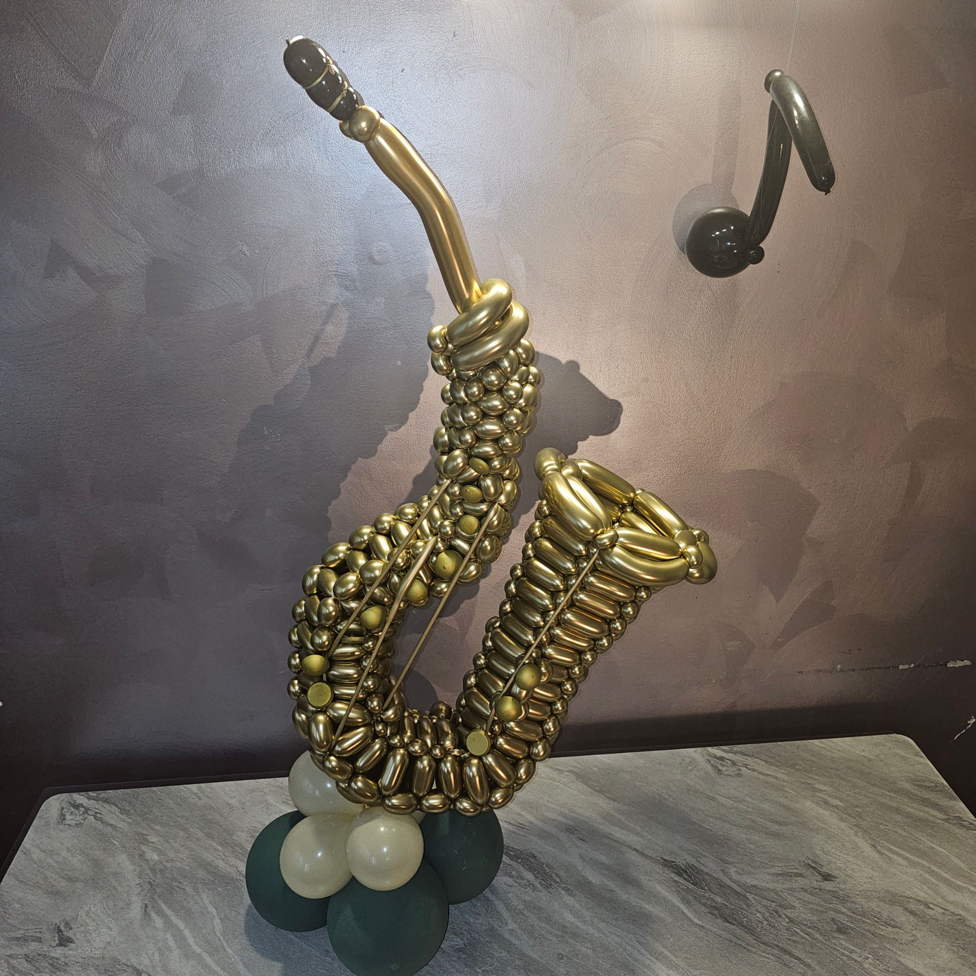 Golden balloon saxophone with musical note, resting on a base of green and white balloons against a gray background.
