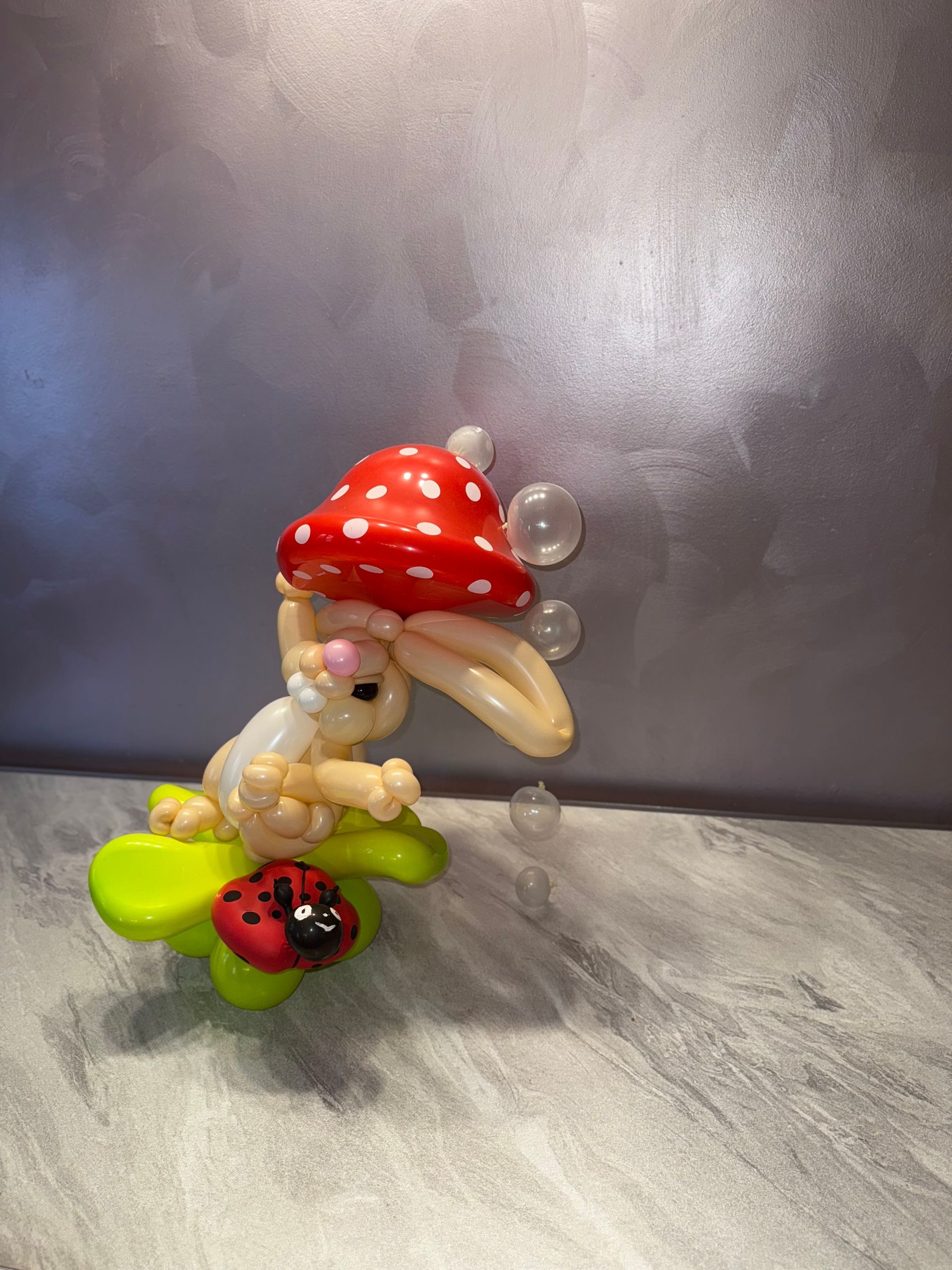 Balloon art: white bunny with red polka dot mushroom hat, on green leaf, with ladybug, bubbles.