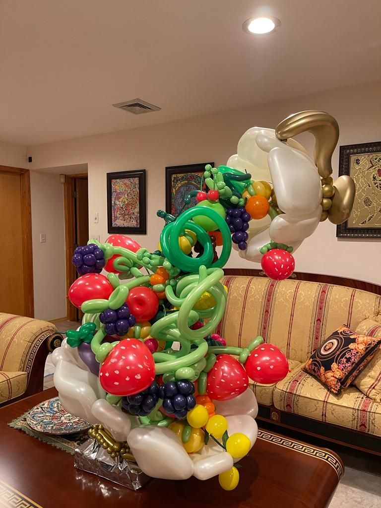 Balloon sculpture of a hand holding a fruit basket, various colorful fruits, indoor setting.