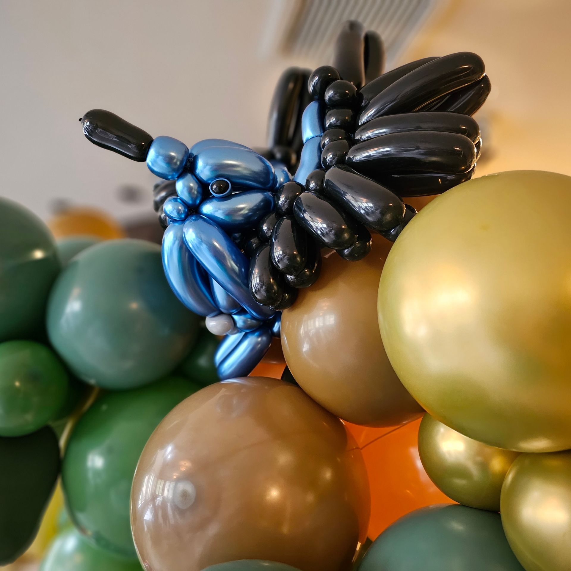 A blue and black balloon dragonfly sculpture on a wall of green, gold, and brown balloons.
