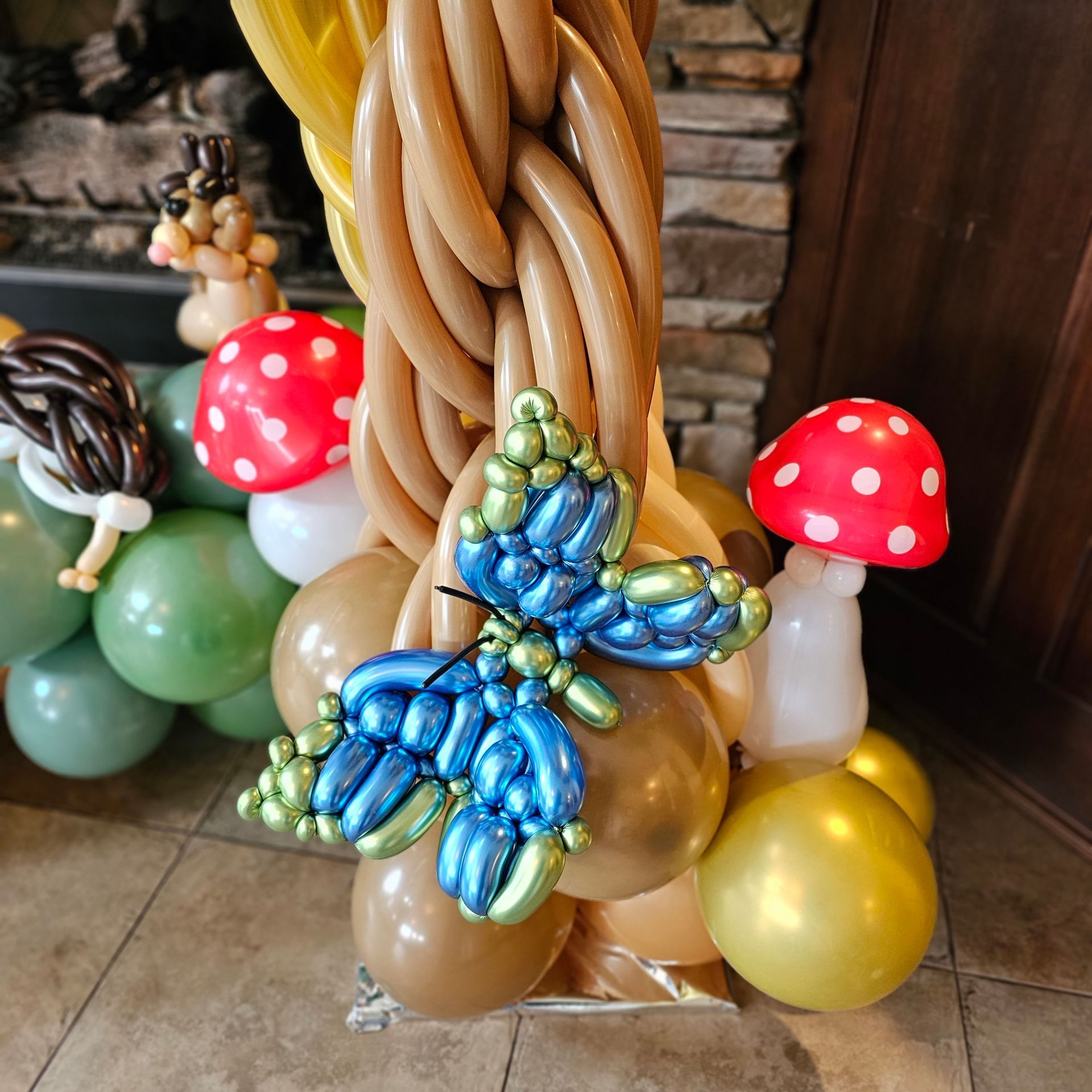 Balloon art of a tree trunk with a blue butterfly, mushrooms, and woodland creatures.