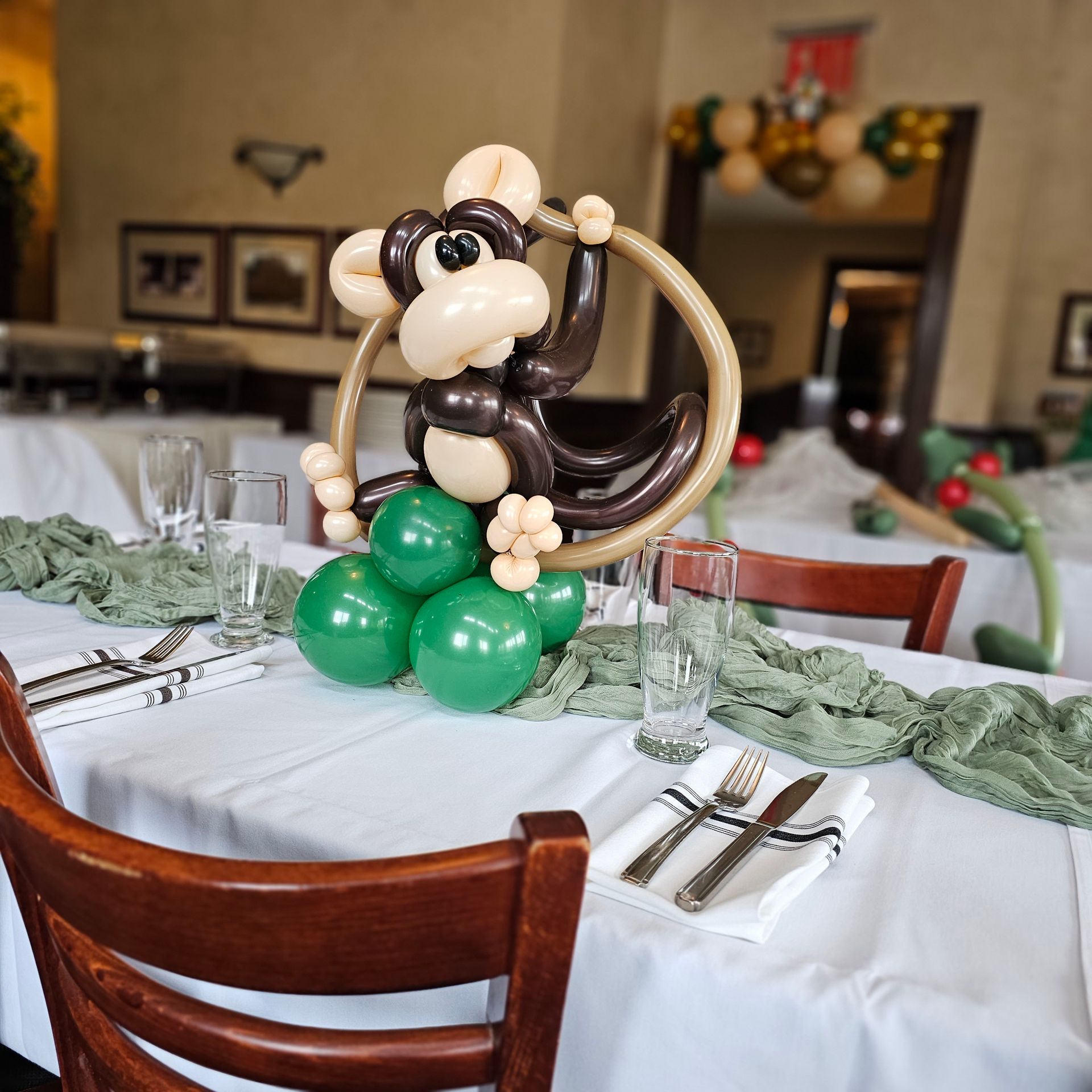 Monkey balloon sculpture centerpiece at a table setting; green balloons, white tablecloth.