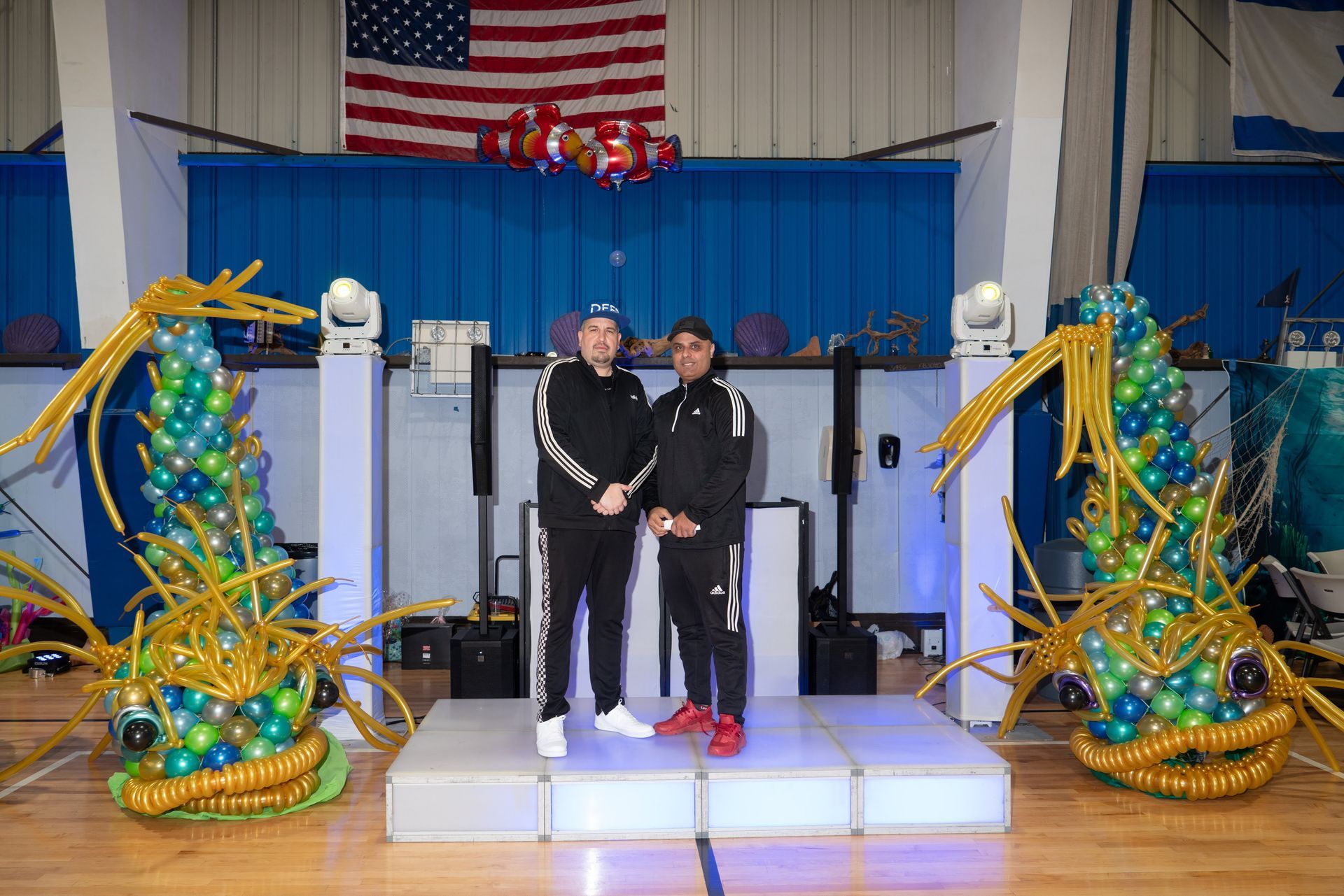 Two men on a stage with balloon fish decorations, in front of an American flag in a gymnasium.