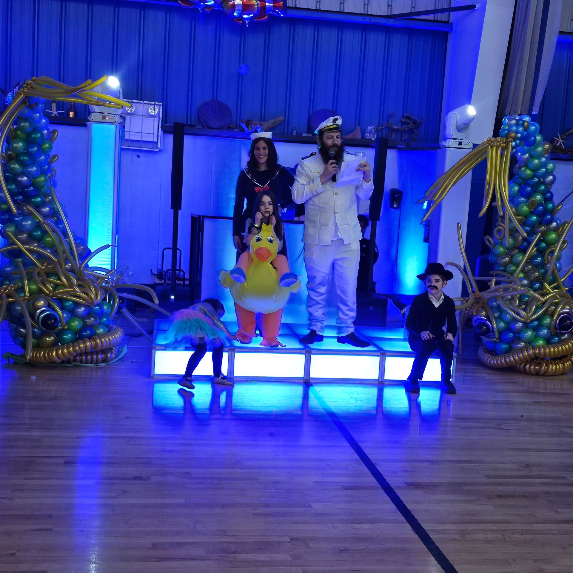 People on stage, one in a sailor costume, with balloon decorations and blue lighting.