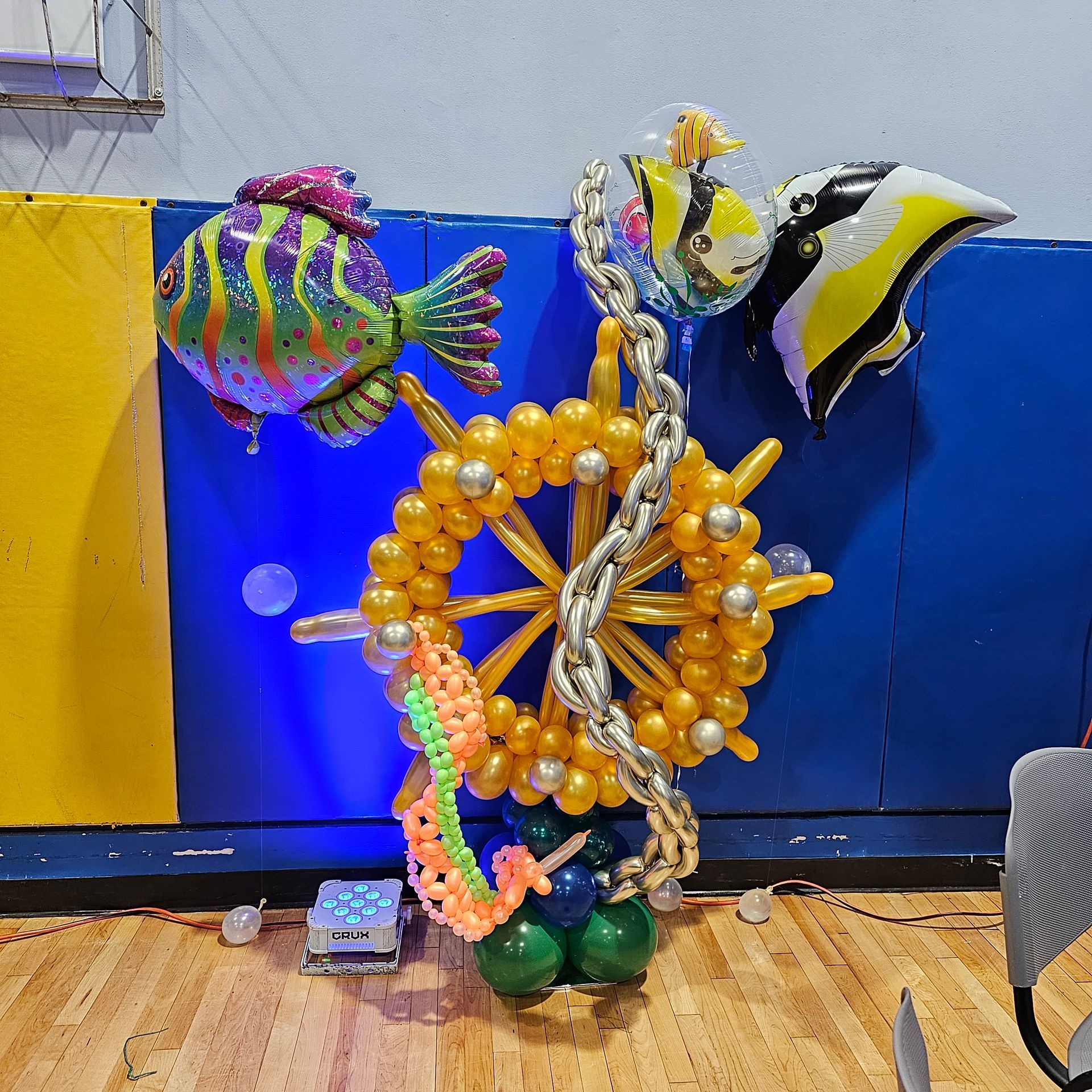 Balloon sculpture of a ship's wheel with fish balloons, on a wooden floor, with a blue and yellow wall background.