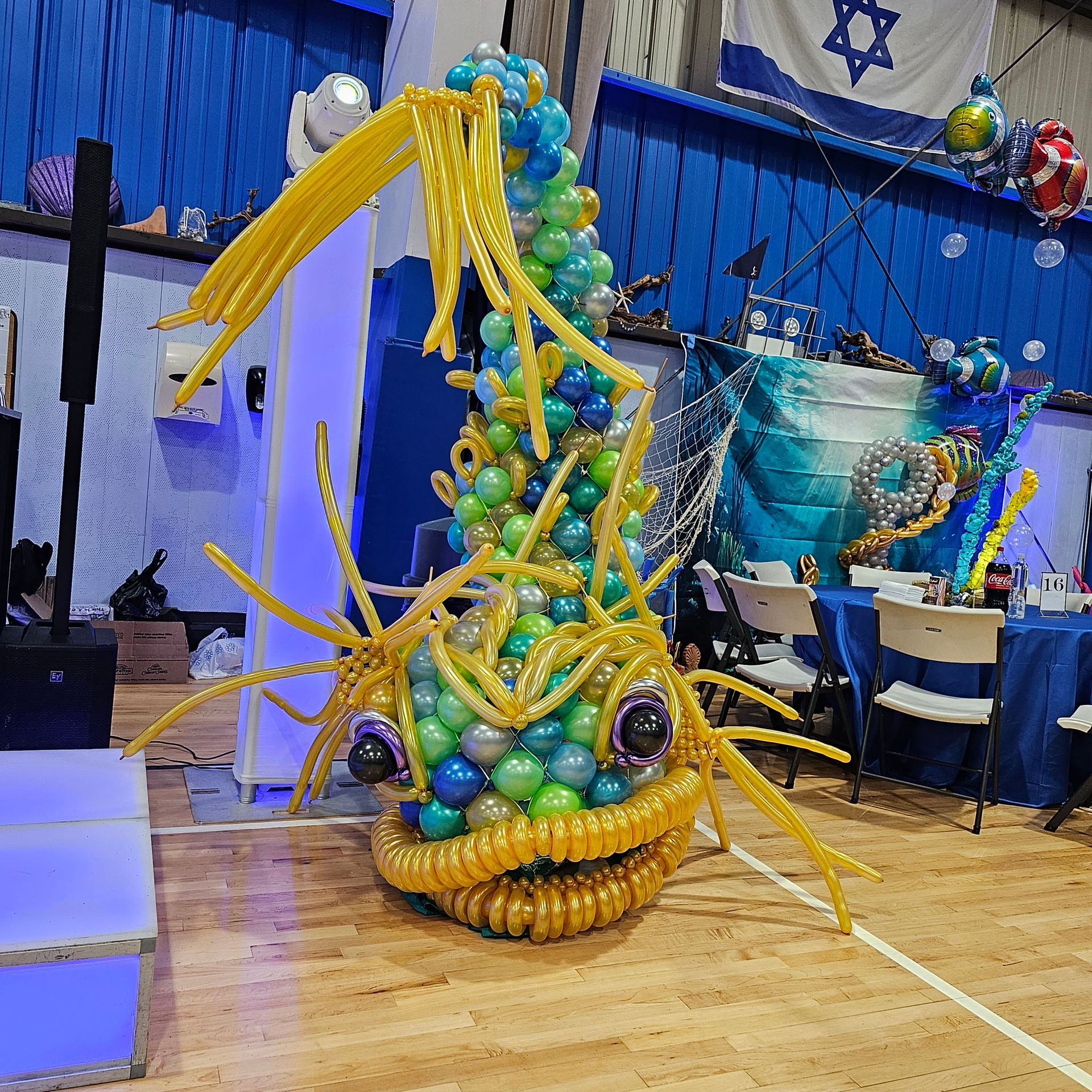 A large, colorful balloon fish sculpture at a party, with a blue and gold color scheme and a Star of David flag.
