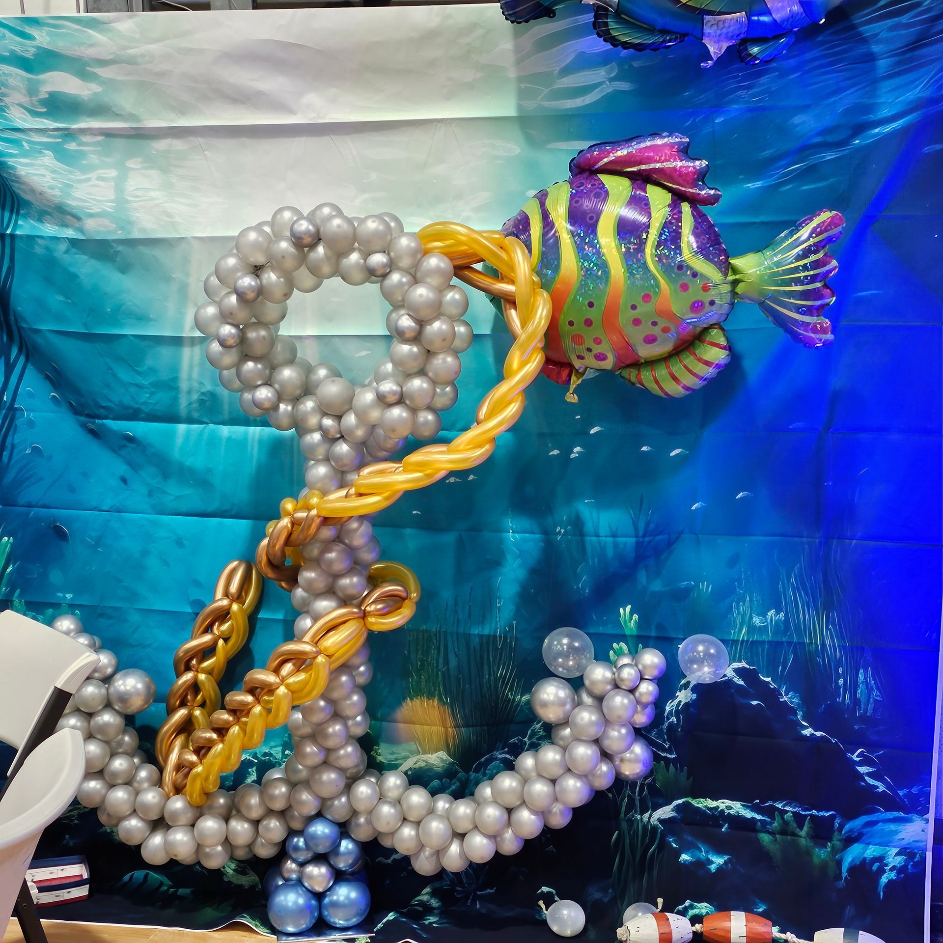 Balloon art: silver anchor with gold rope, colorful fish against an ocean-themed backdrop.