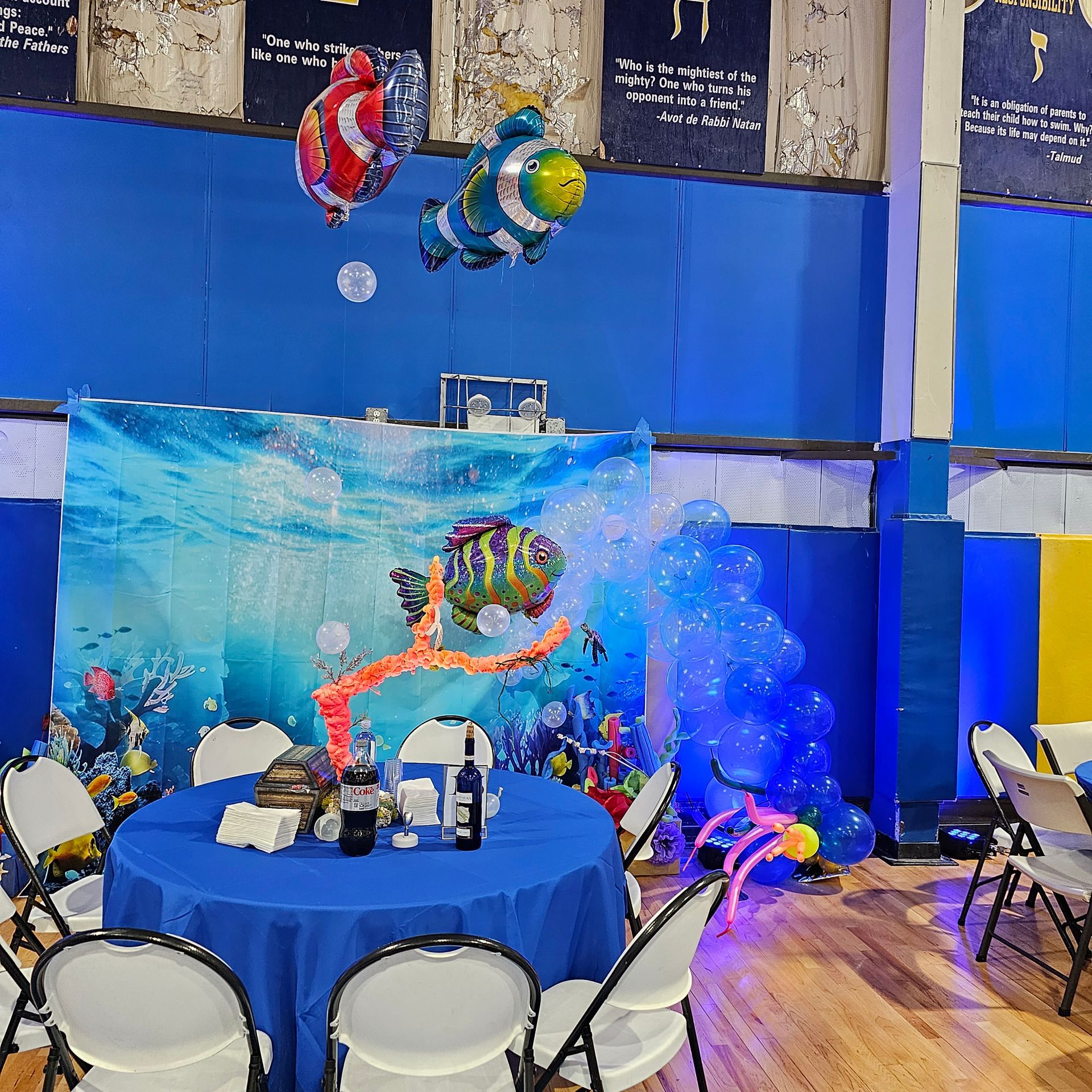 Aquatic-themed party decoration: table with blue cloth, underwater backdrop, clownfish balloons, and chairs.