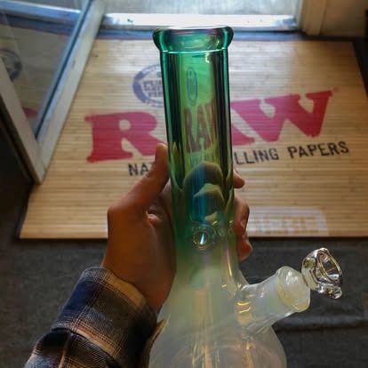 Person holding a green and white glass bong, in front of a RAW rolling papers mat.