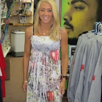 Woman in a patterned dress smiles in a shop; clothing racks and merchandise visible.