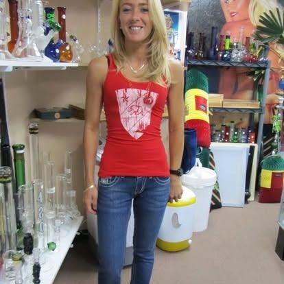 Woman in red tank top and jeans smiles in a smoke shop, surrounded by glass pipes.