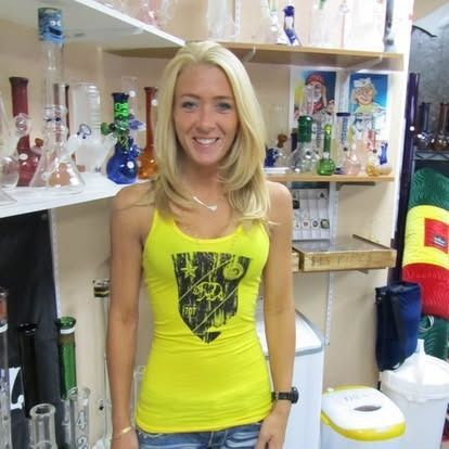 Woman in yellow tank top smiling, surrounded by glass smoking pipes on shelves.