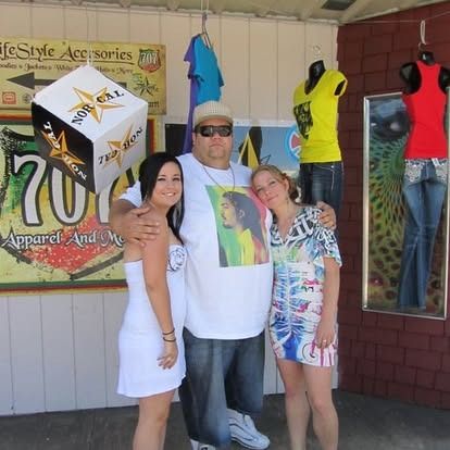 Three people pose in front of a storefront: a man with sunglasses and two women. Storefront displays clothing and a large cube sign.