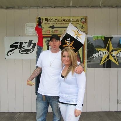 Man with arm around woman, posing in front of store signs, holding a die.