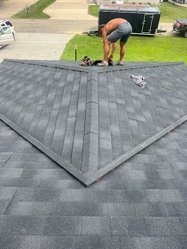 A person in shorts on a dark grey shingled roof, setting down shoes near a triangular section. Green grass and vehicles are in the background.