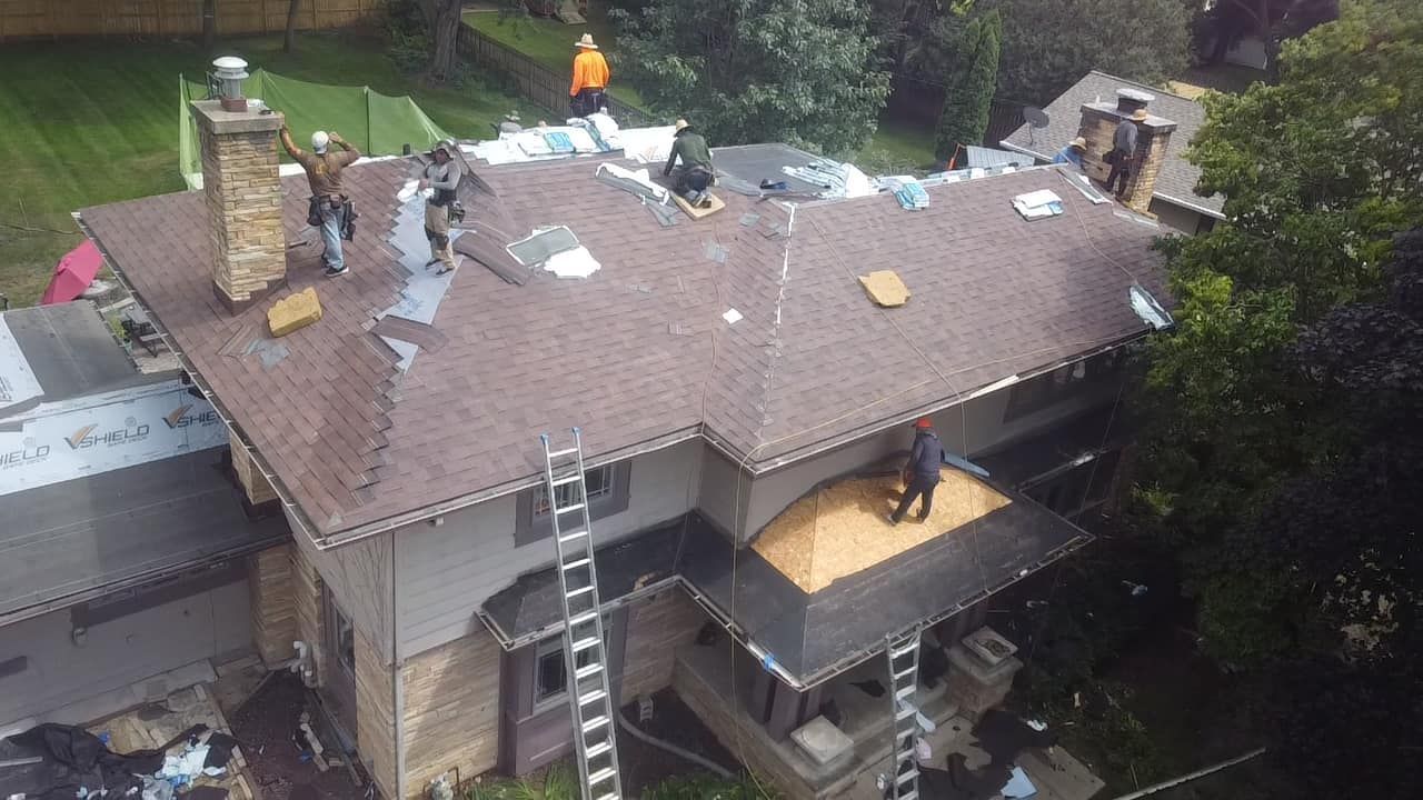 Roofers working on a house roof, likely installing shingles. Ladders and a green lawn are visible in the setting.