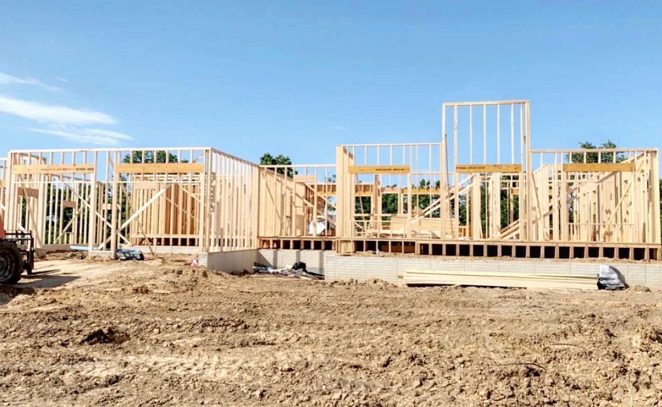 Construction site with a wood-framed house in progress against a blue sky. The foundation is in place, and walls are being built.