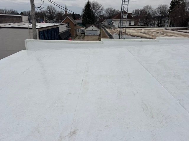 View from a flat, white commercial roof overlooking a town with buildings and trees. The sky is overcast.