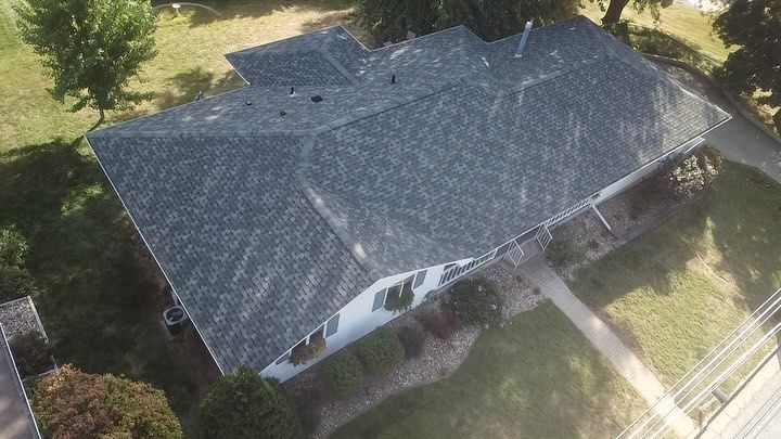 Aerial view of a house with a dark gray shingled roof, surrounded by green grass and a few trees.