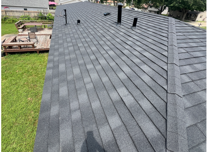 Newly shingled roof with shades of gray shingles, black vents and pipes, and a wooden deck with furniture in the yard.