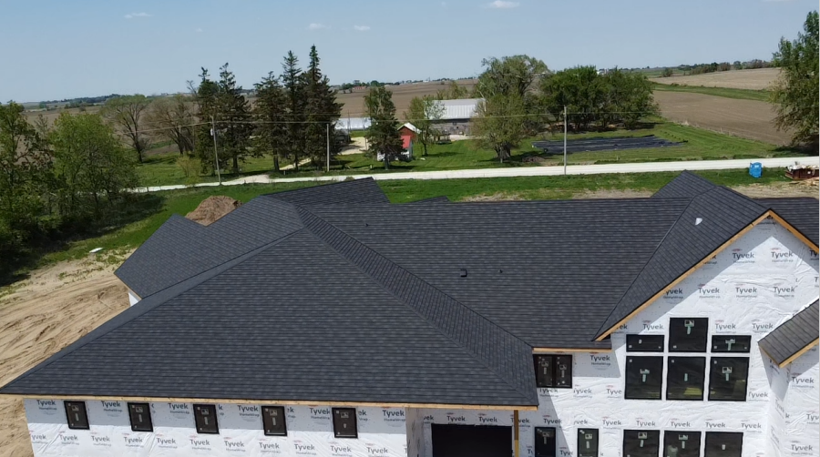 A partially constructed house with a dark gray roof and wrap, set against a rural backdrop.