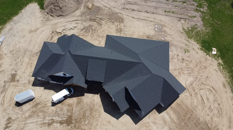 Aerial view of a dark gray shingled roof on a house under construction. White work vehicles and a sand-filled lot surround the house.
