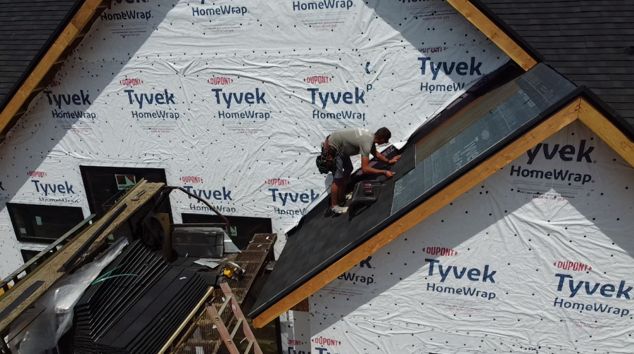 Two roofers installing shingles on a new roof, with Tyvek house wrap visible.