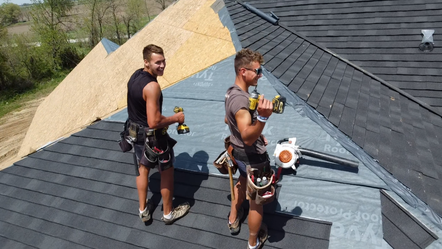 Two roofers installing shingles on a house roof; one smiles at the camera, both hold drills, sunny day.