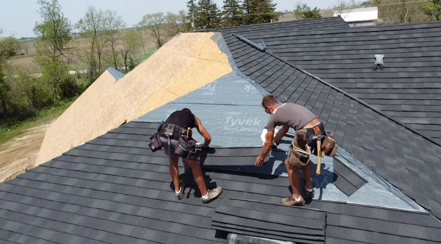Two roofers installing asphalt shingles on a house roof; one is hammering, and the other is holding shingles.