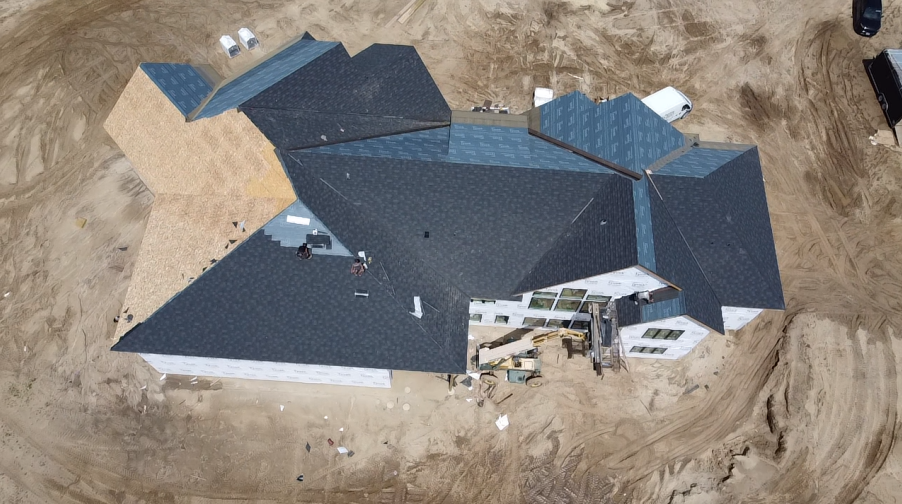 Aerial view of a house under construction with a complex, dark gray roof and unfinished light wood sections. The setting is a dirt lot.