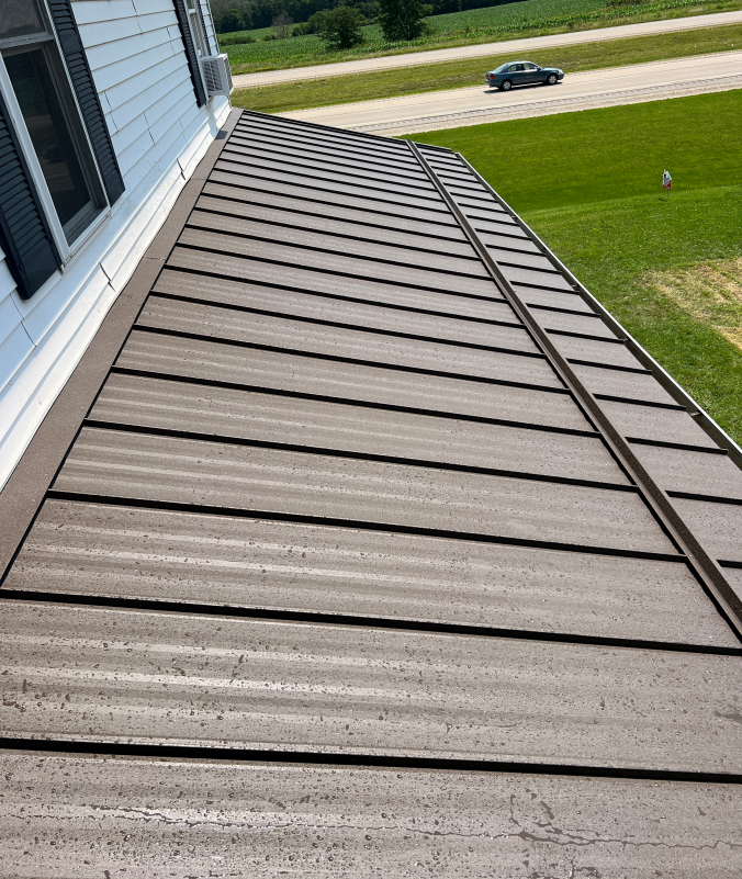 Brown metal roof on a white house, seen from above. Green grass and road in the background.