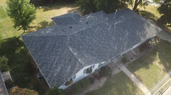 Overhead view of a house with a dark gray shingle roof, surrounded by green grass and a few trees.