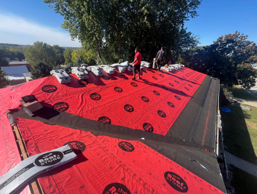 Two roofers on a partially-shingled roof, covered in red underlayment. They're installing new shingles on a sunny day.