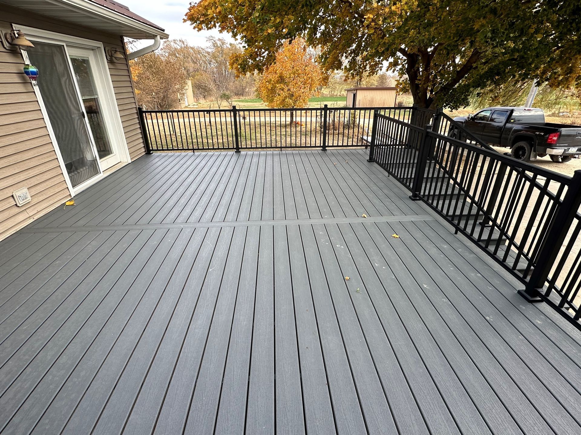 Grey composite deck with black railing attached to a light brown house. A black truck is parked nearby under a tree with fall foliage.