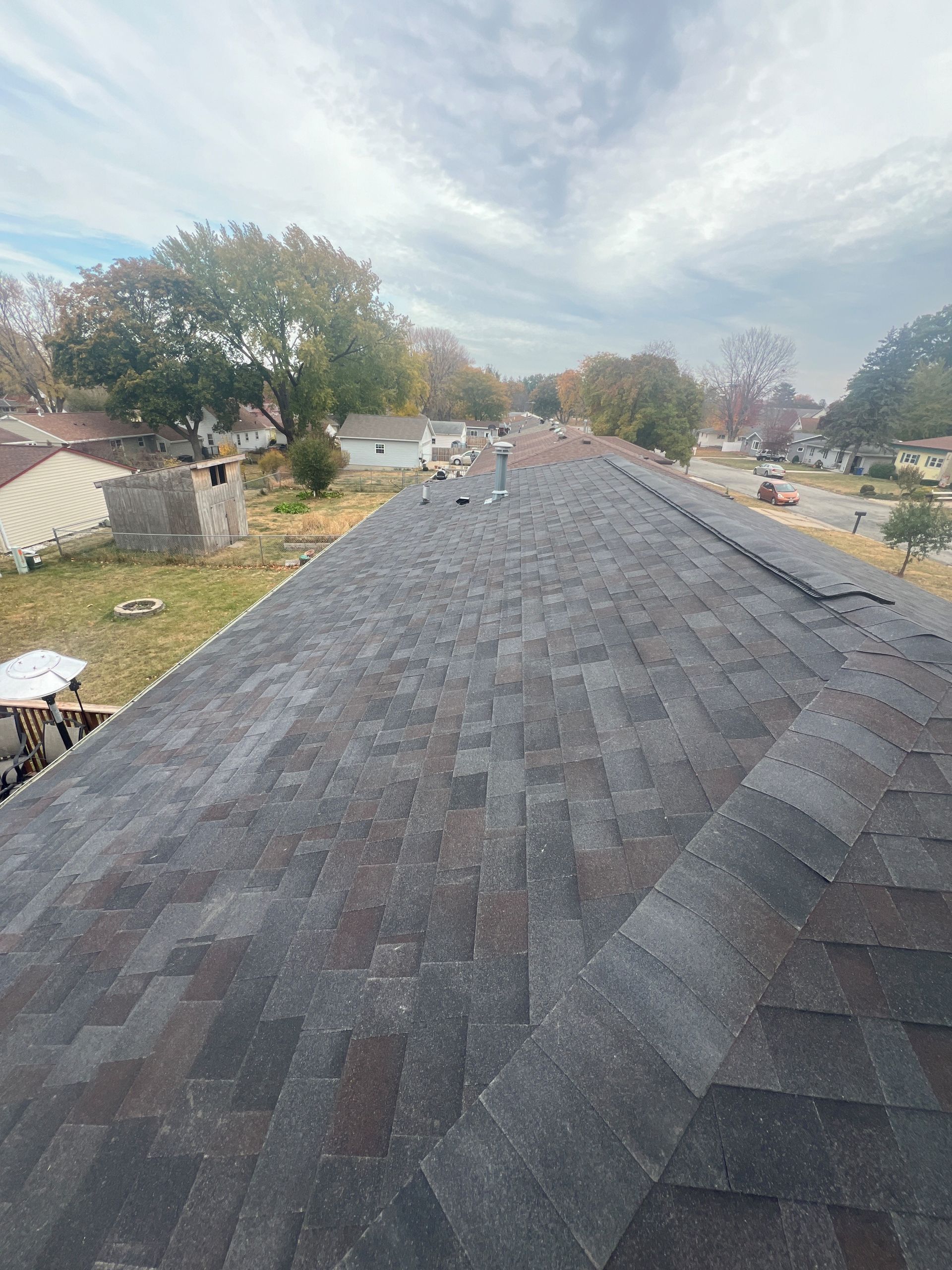 View from a roof showing asphalt shingles, a neighborhood in the distance, and a cloudy sky.