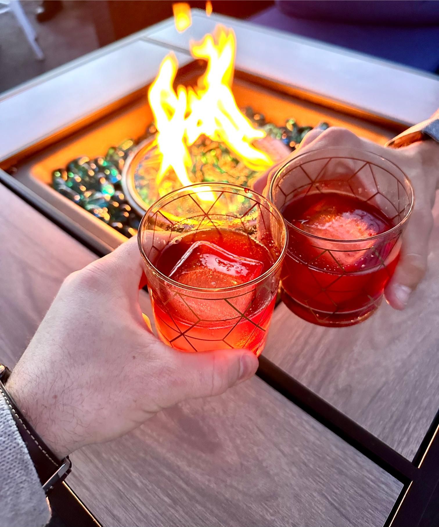 A person is holding two glasses of red liquid in front of a fire pit