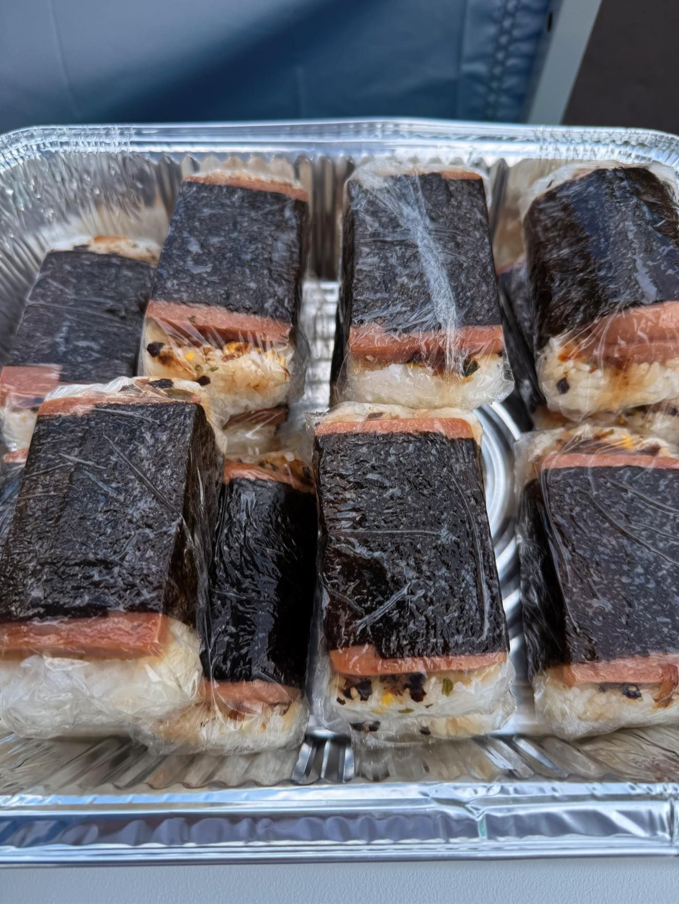Spam musubi in a foil tray, wrapped in plastic. Square rice with grilled spam, wrapped in nori seaweed.