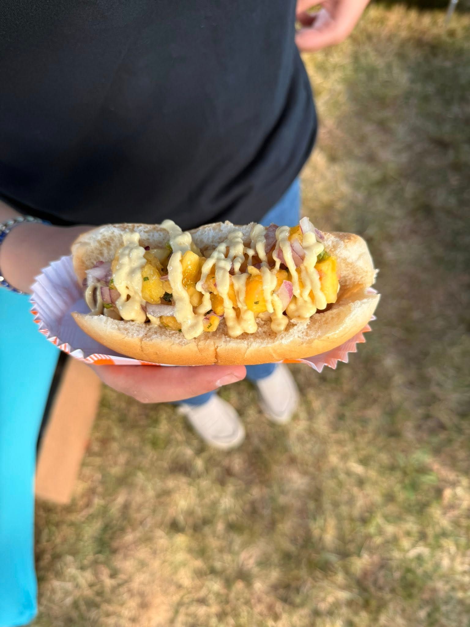 Hot dog in bun topped with yellow relish and sauce, held by a person outdoors.