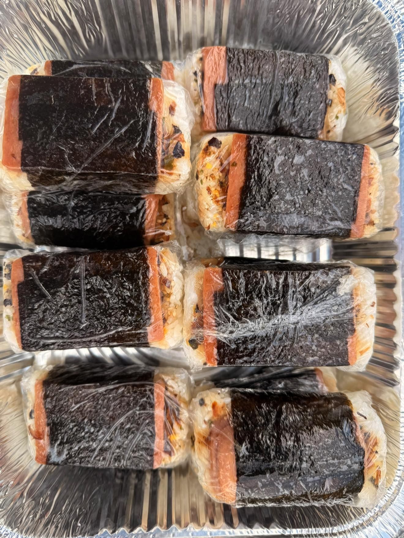Spam musubi wrapped in plastic, in a foil container. Rectangular rice with cooked spam, and nori.
