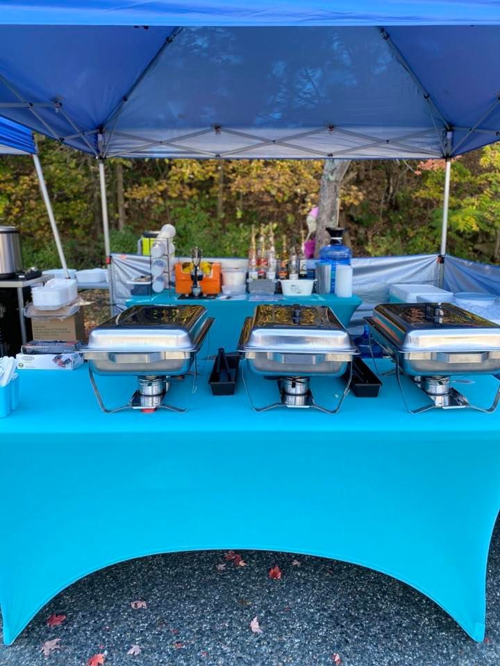 Outdoor catering setup under a blue canopy with three covered chafing dishes on a teal table.