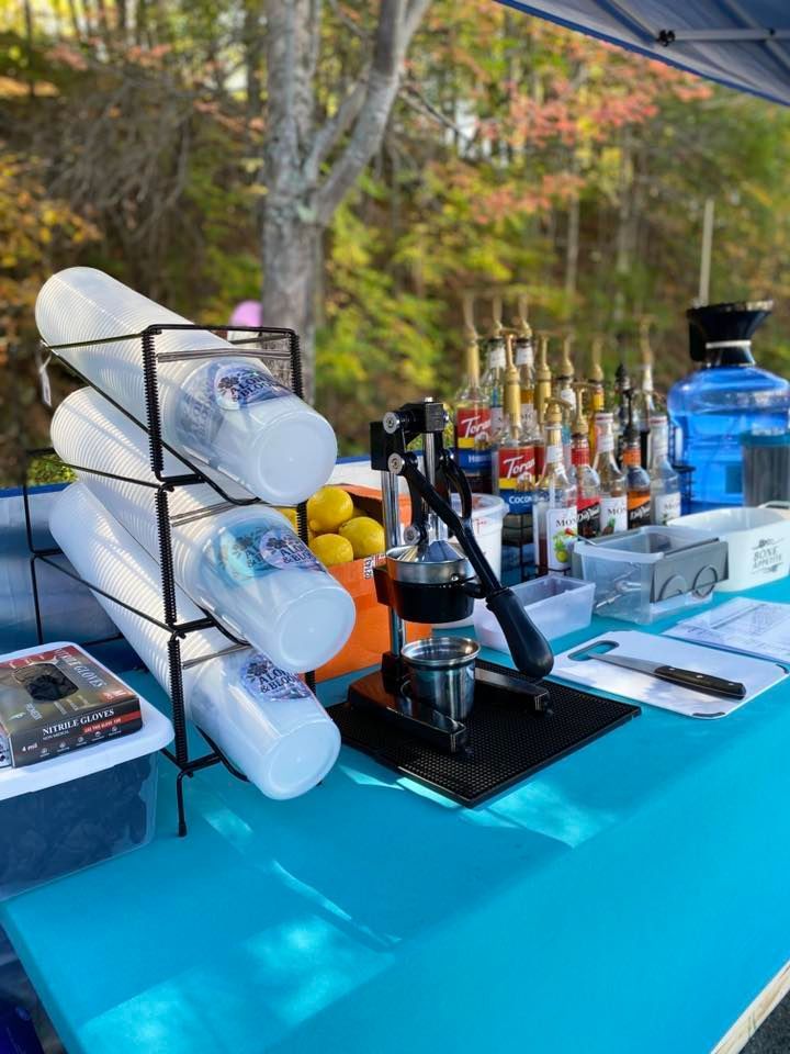 A blue table with a drink station, including cups, syrups, and a citrus press. Outdoors.