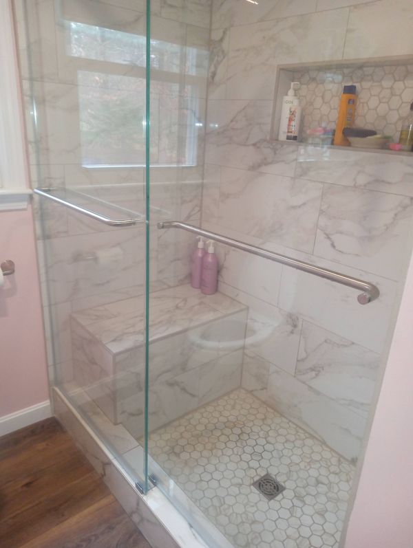Glass-enclosed shower with marble-look tile, built-in seat, and grab bars.
