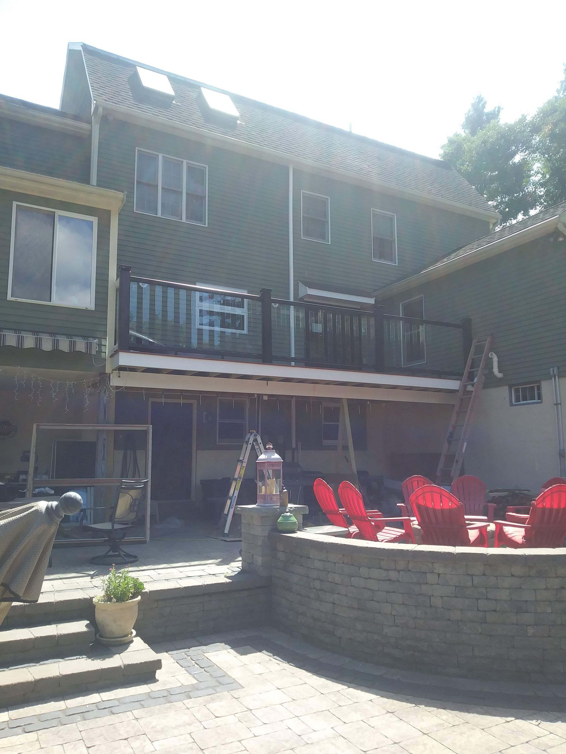Back of a two-story green house with a deck being built over a patio with red chairs and a fire pit.
