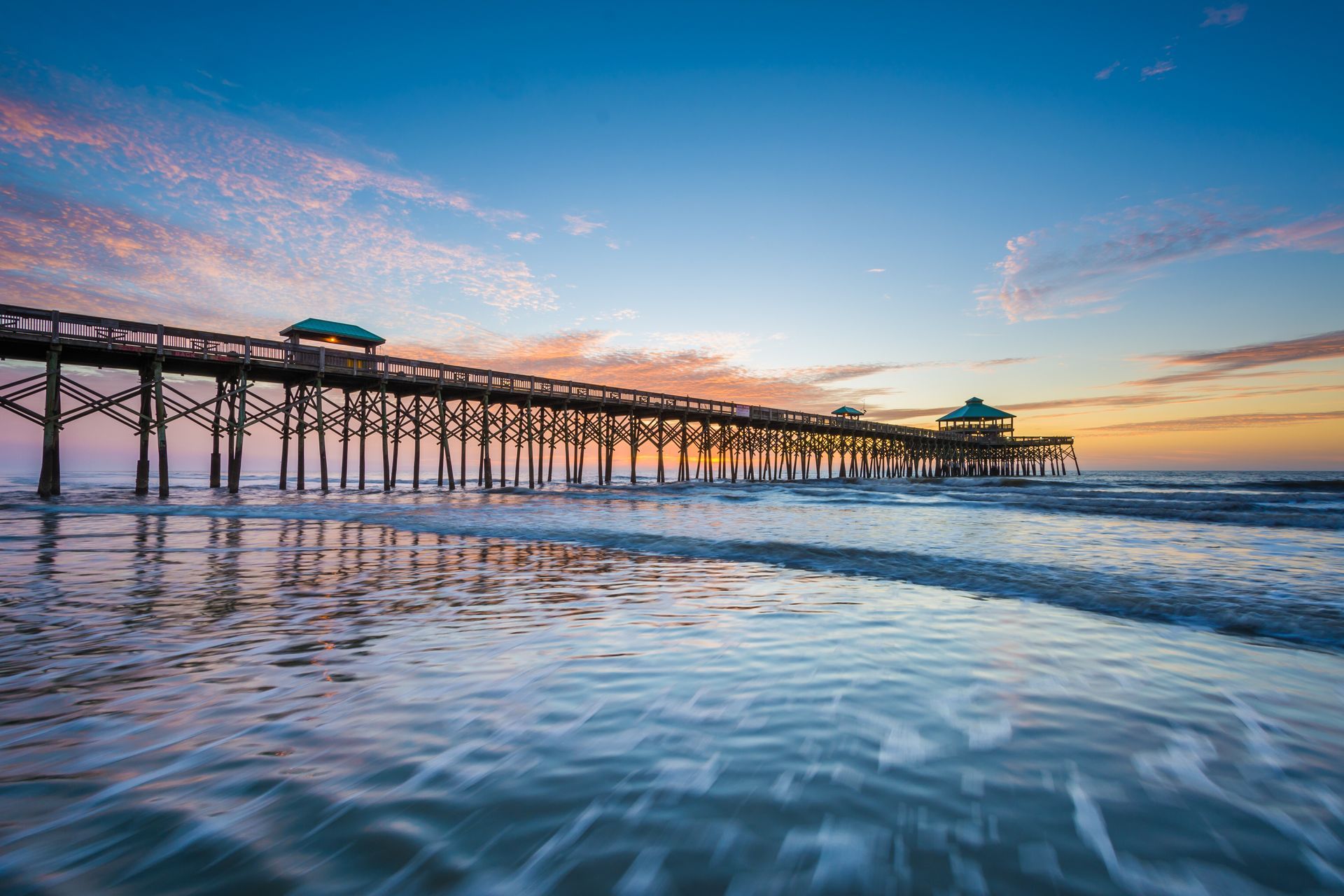 Wooden pier extending into ocean at sunset; pink and orange sky, blue water.