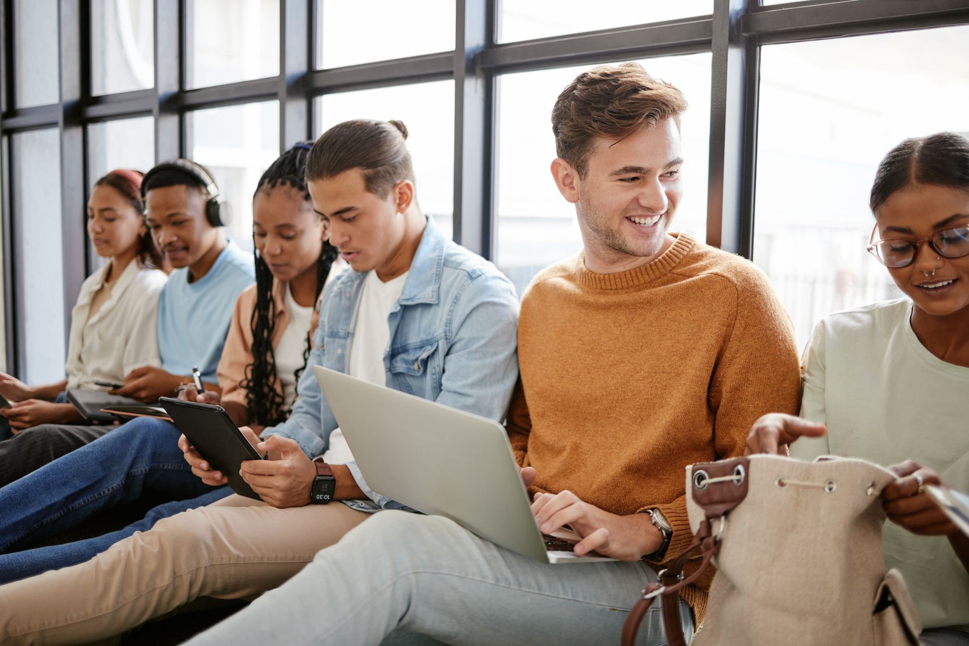 A group of young people are sitting in front of a window using laptops and tablets.